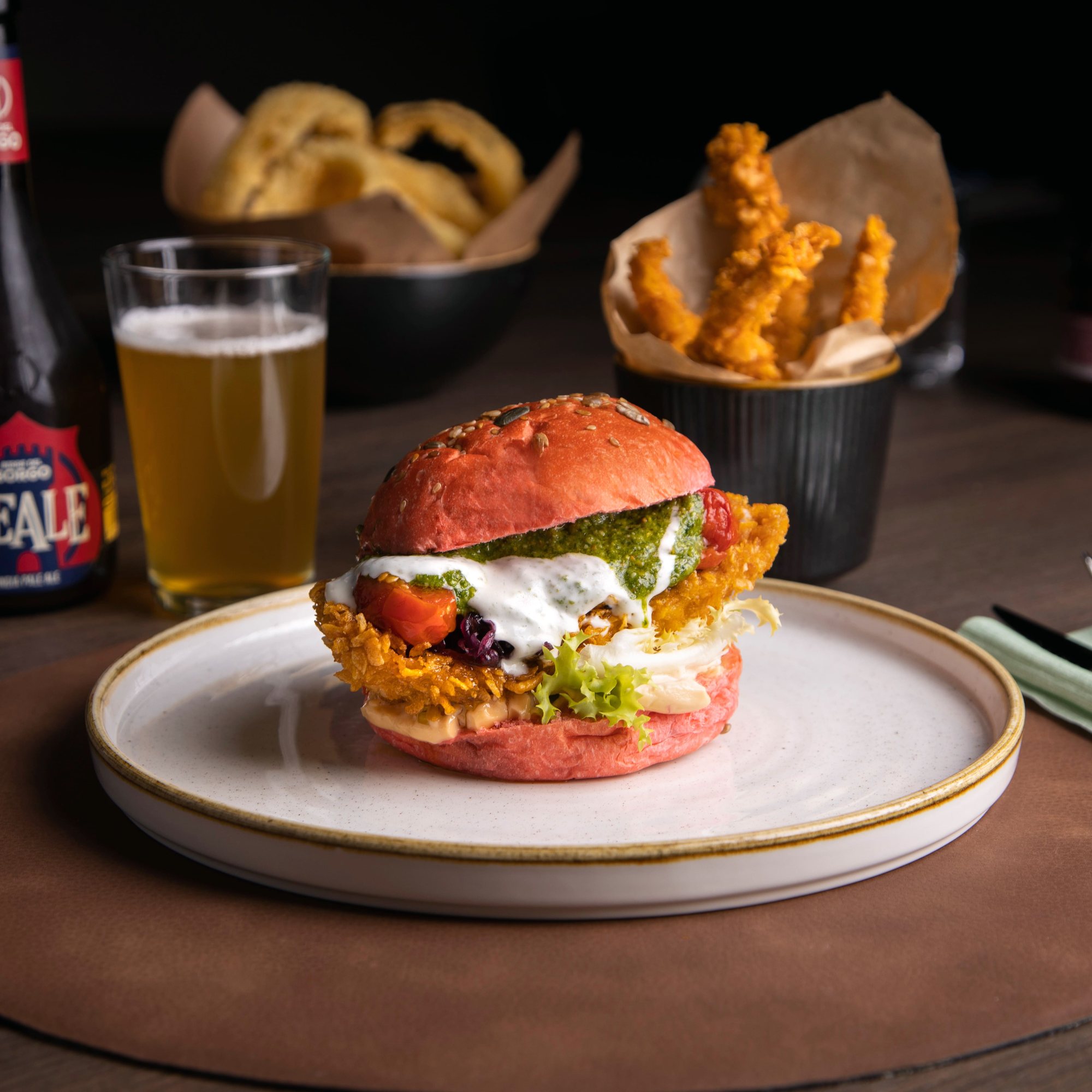 Burger con bun rosso, pollo croccante e pesto, con una birra sul tavolo