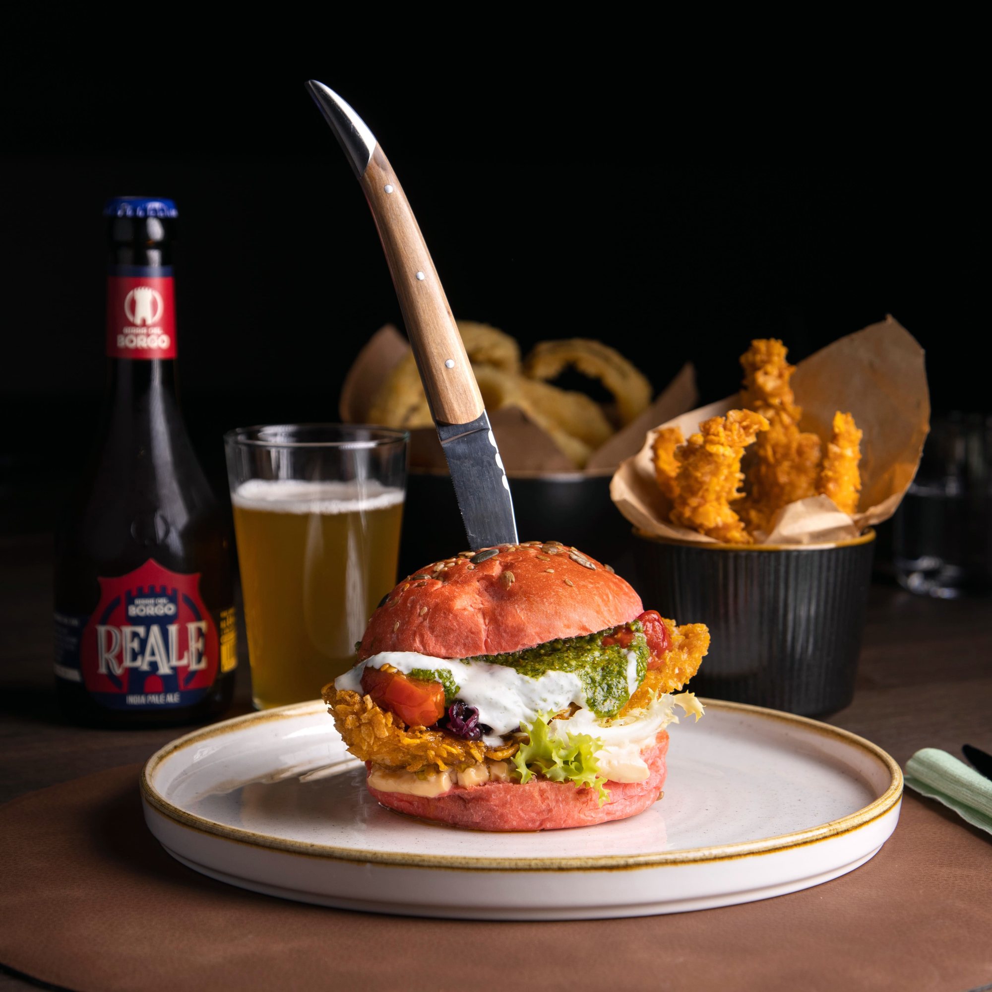 Burger con bun rosso, birra Reale, onion rings e patatine su un tavolo del locale